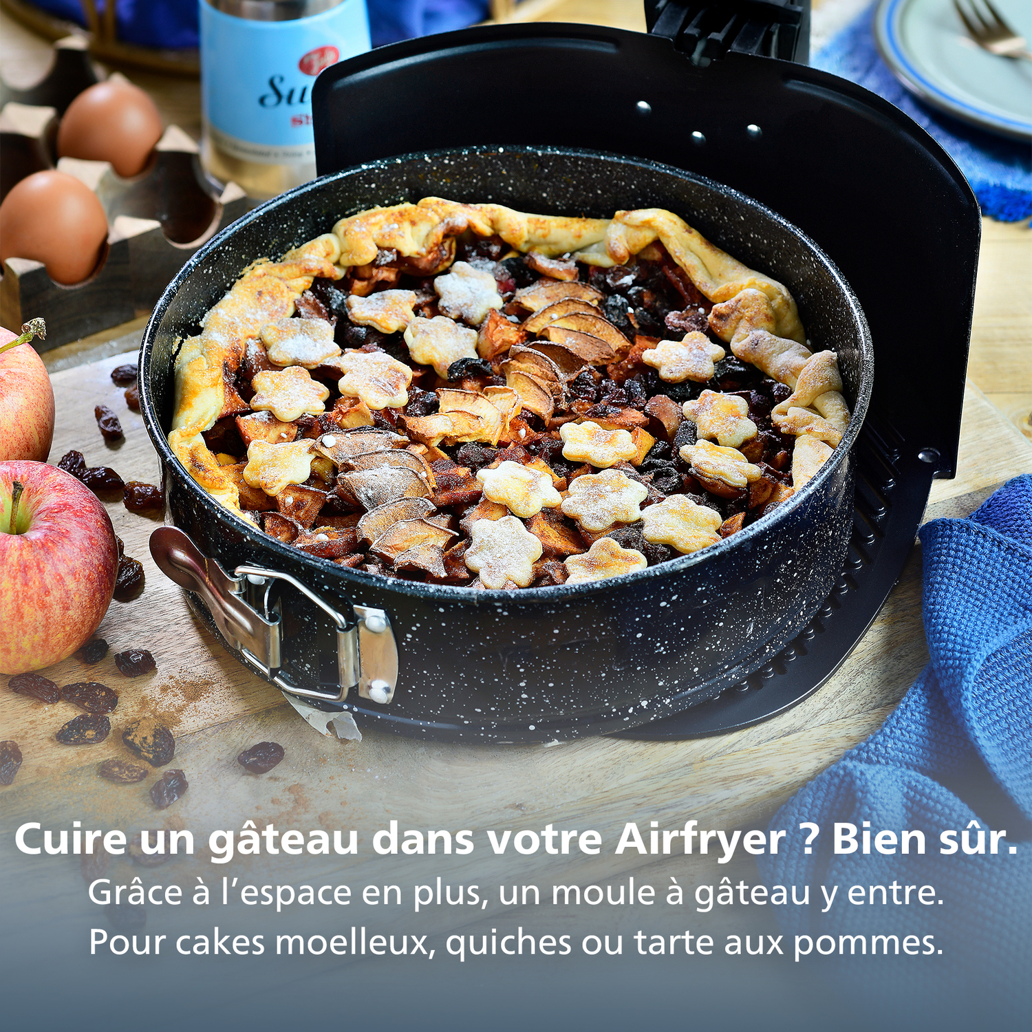 Tarte aux pommes dans un moule noir. Pommes et raisins secs éparpillés. Œufs et bouteille en arrière-plan.