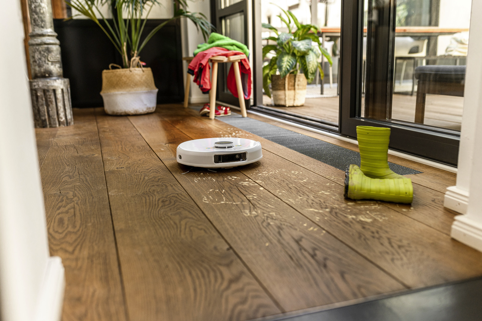 Egy fehér robotporszívó egy fa padlón. Zöld csizmák, növények és egy ruhákkal teli zsámoly látható a szobában.
