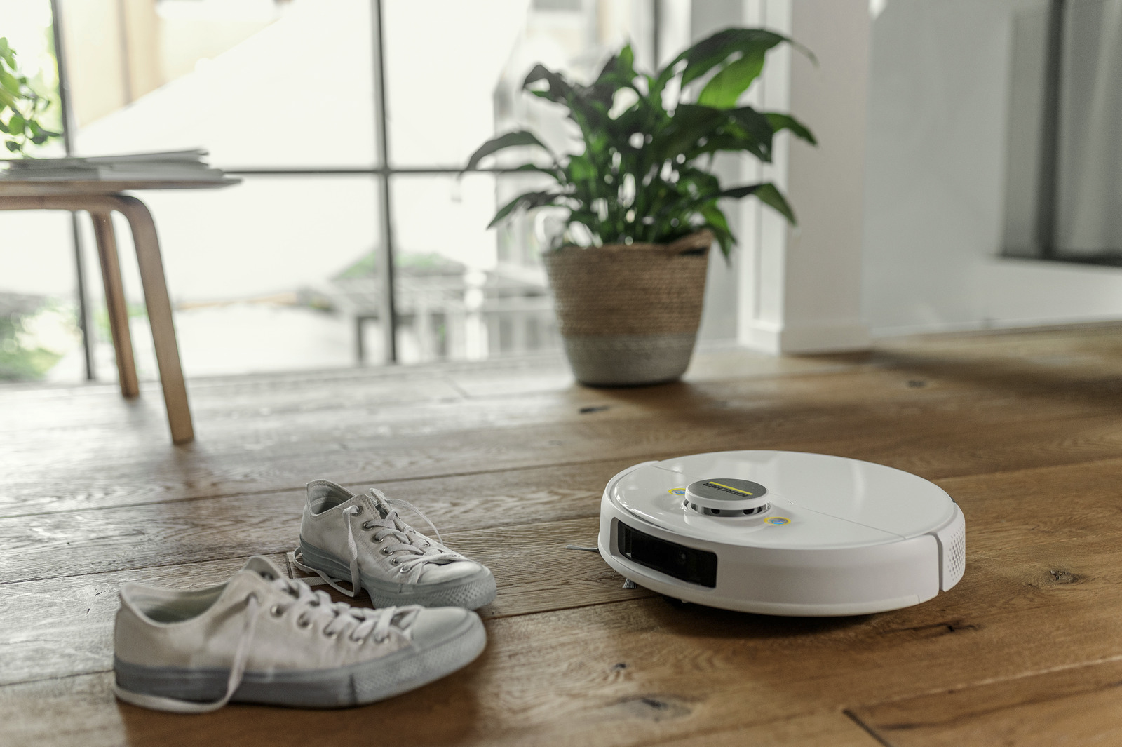 Egy fehér robotporszívó, szürke tornacipők és egy cserepes növény a fa padlón. A háttérben egy asztal látható.