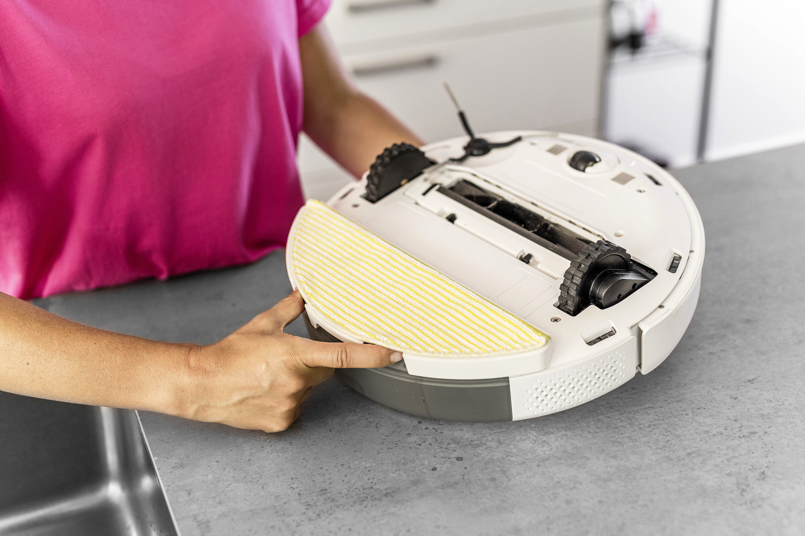 Egy személy egy fehér robotporszívót tart. A tisztítóhoz egy sárga-fehér felmosófej van rögzítve, és egy rózsaszín ing látható.