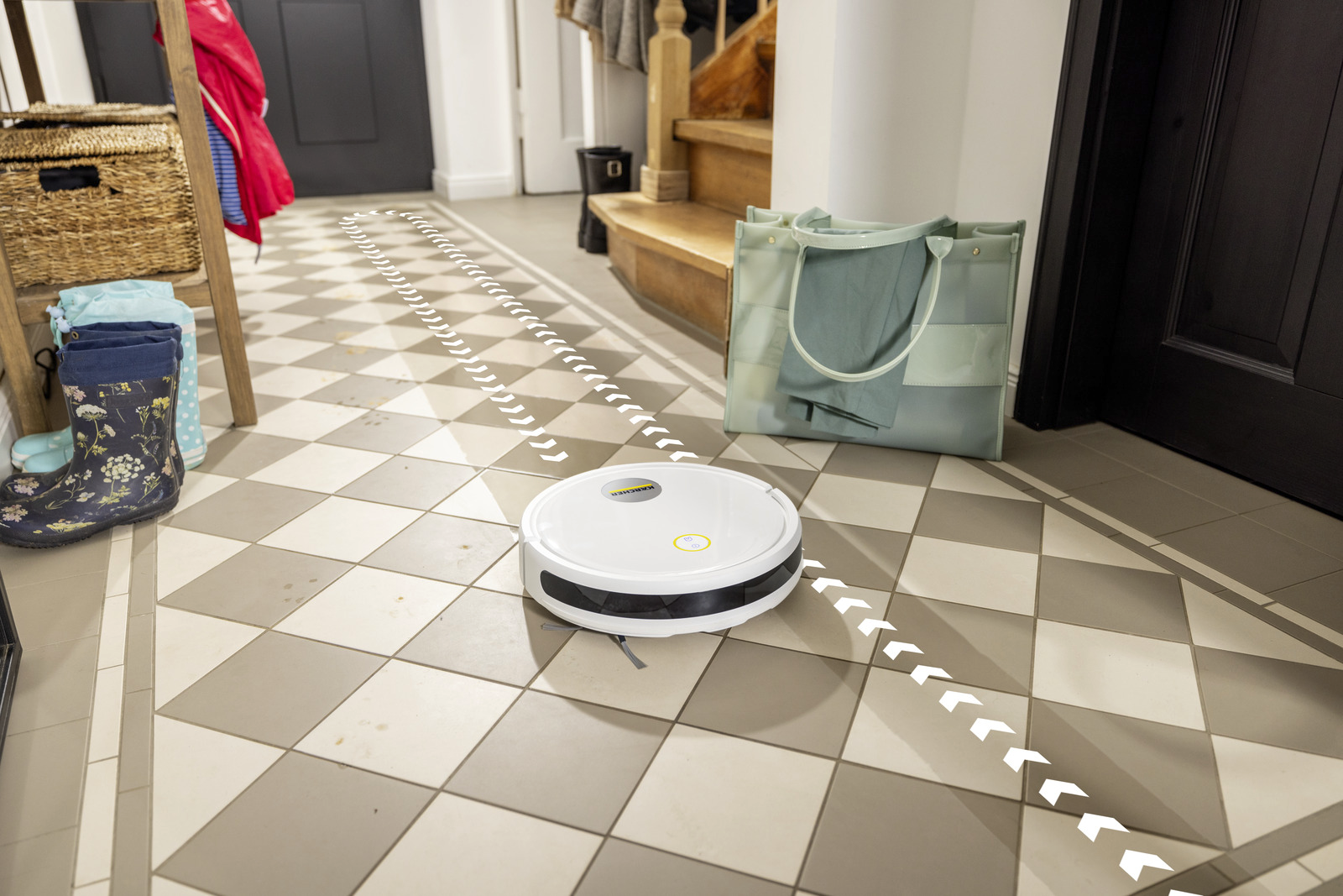 Egy fehér robotporszívó takarít egy kockás padlót. Egy táska, csizmák és egy fa állvány láthatók a háttérben.