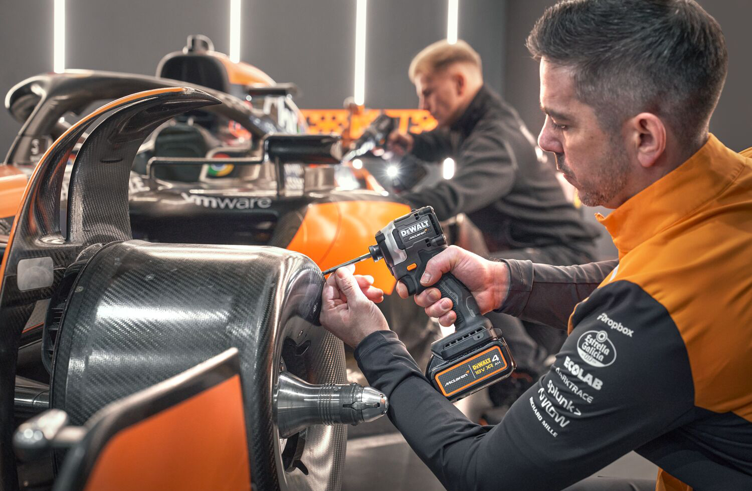 Egy szerelő elektromos fúrót használ egy McLaren versenyautón. Az autó narancssárga és fekete. Egy másik szerelő dolgozik a háttérben.