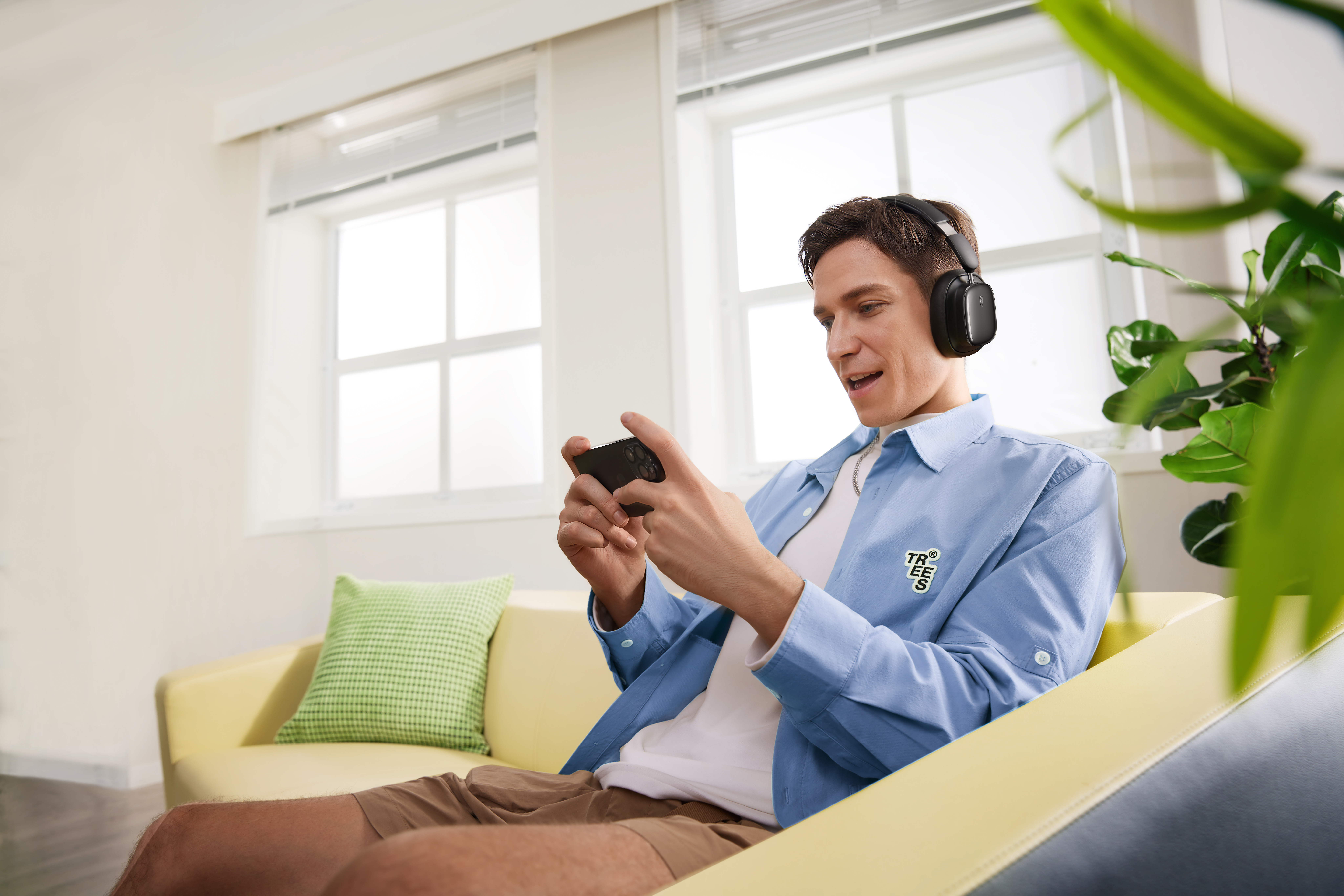 Egy férfi fejhallgatóval játszik a telefonján. Egy sárga kanapén ül. Van egy zöld párna és egy növény.