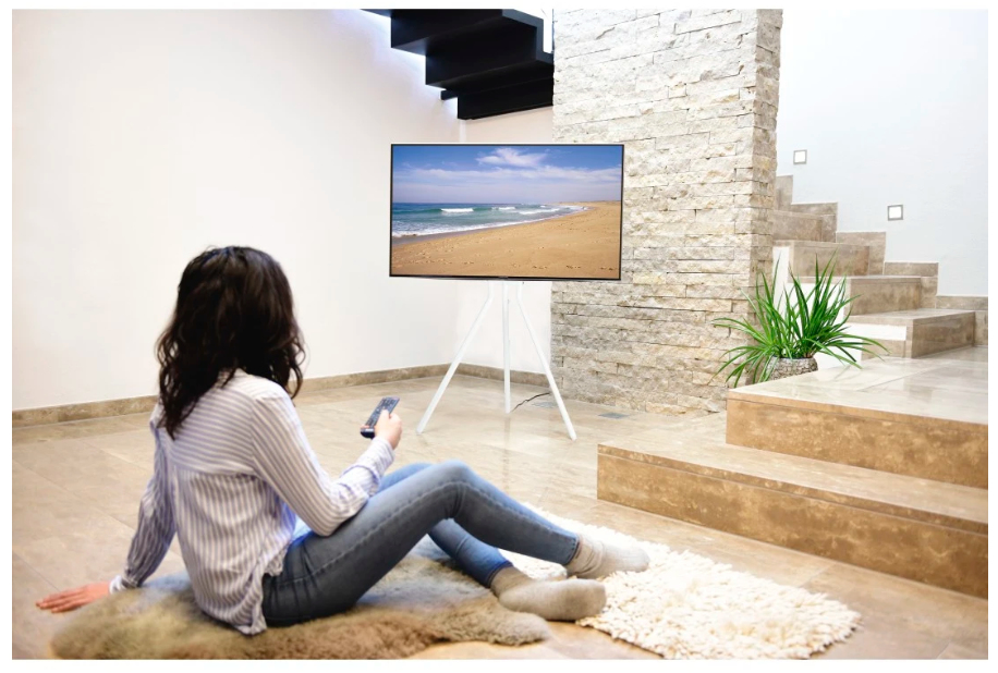 Una mujer sentada en una alfombra ve la televisión, que muestra una playa. La TV está en un trípode blanco. Escaleras y una pared de piedra al fondo.