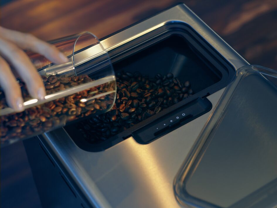 Una mano vierte granos de café de un recipiente de vidrio a un compartimento de la cafetera. Los granos son marrones.