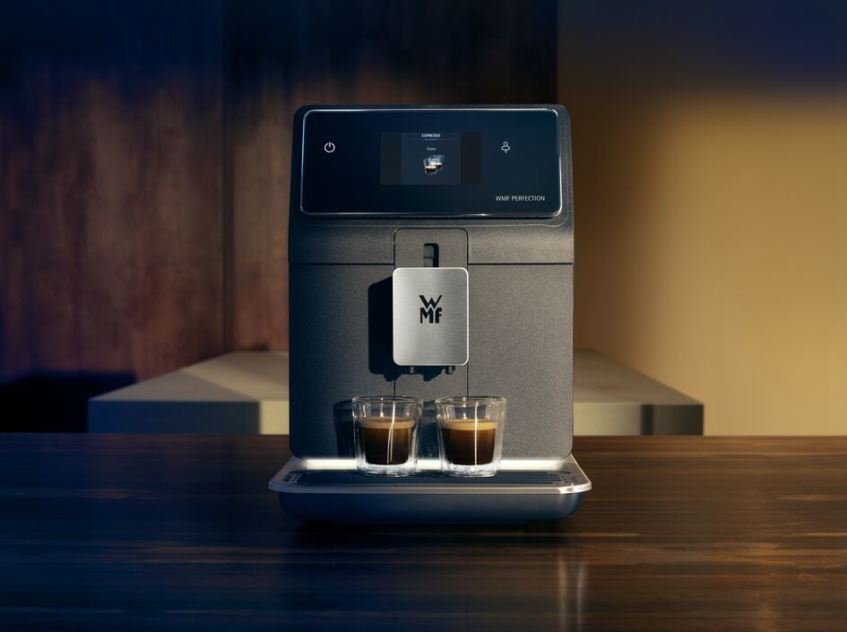 Una cafetera con dos chupitos de espresso en vasos transparentes. Tiene una pantalla que muestra 'Espresso'. La máquina es gris oscuro.
