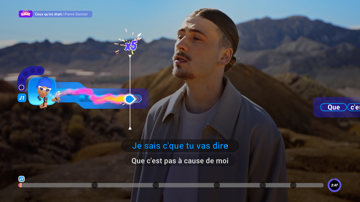 Un uomo canta in un microfono, con testo sullo schermo. Un'interfaccia di gioco è visibile, con un punteggio di x5. Lo sfondo sono montagne sfocate.