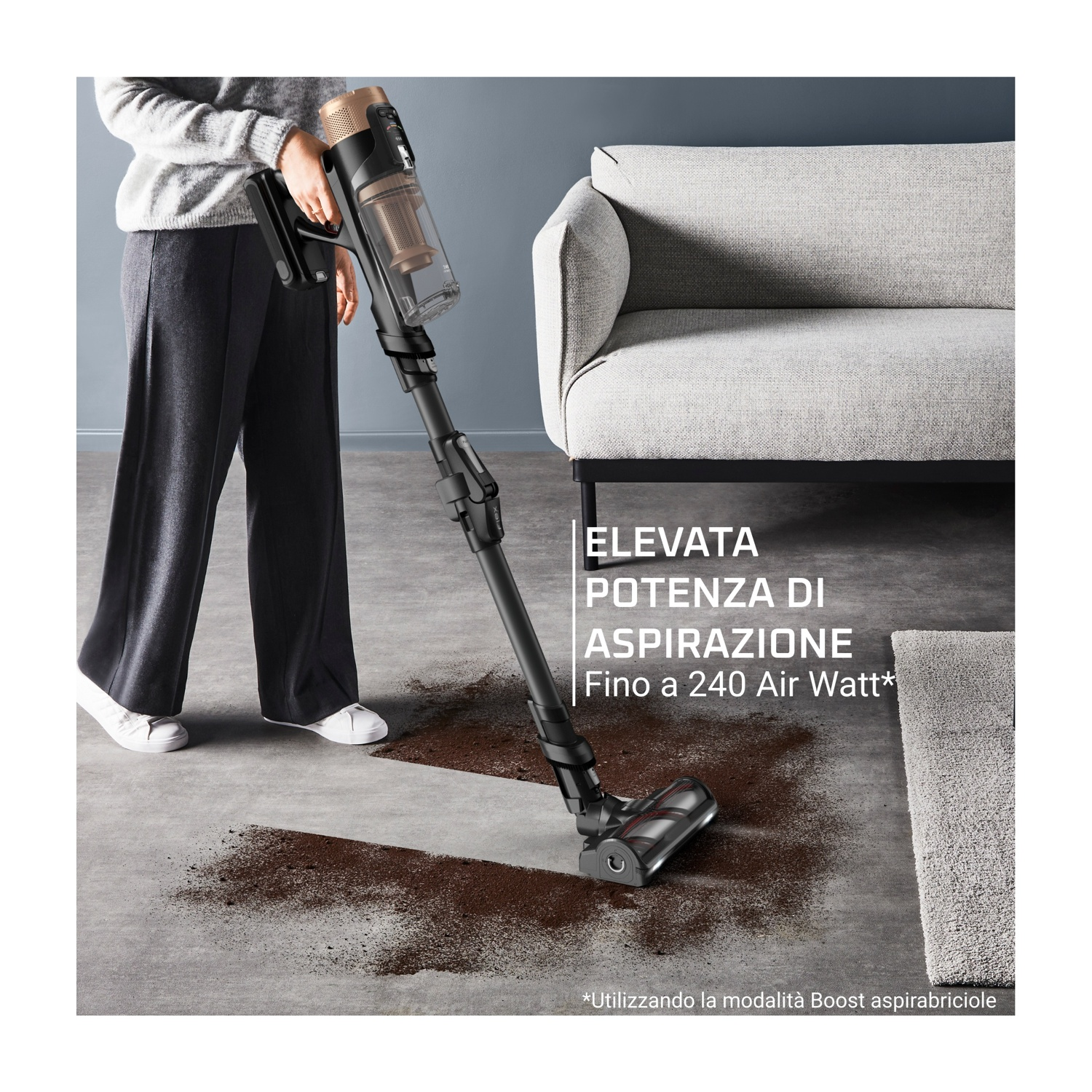 Una persona aspira detriti sul pavimento con un aspirapolvere nero e bronzo. Un divano e un tappeto sono sullo sfondo.