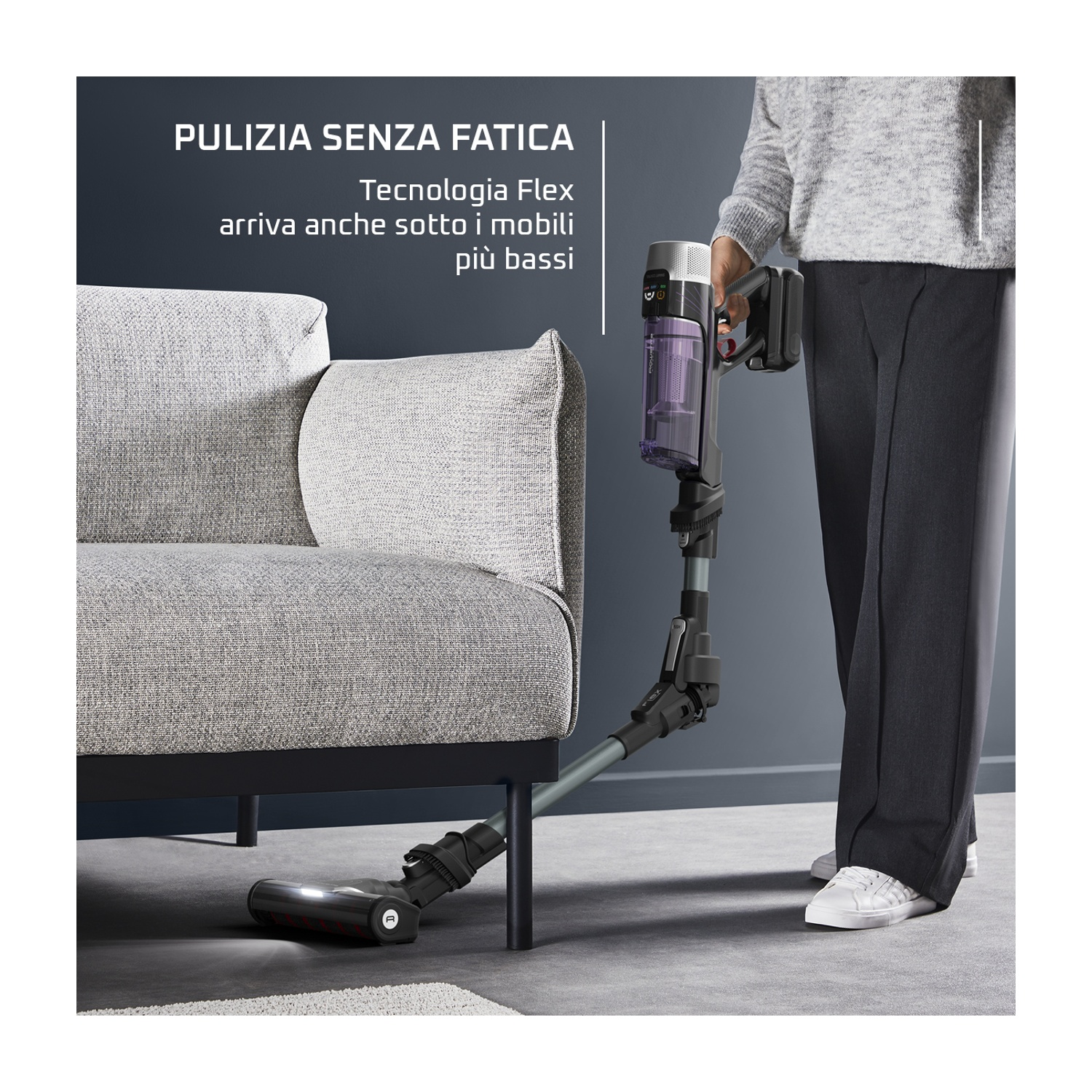 Una donna usa un aspirapolvere sotto un divano. L'aspirapolvere è nero e grigio. Indossa un maglione grigio e pantaloni neri.