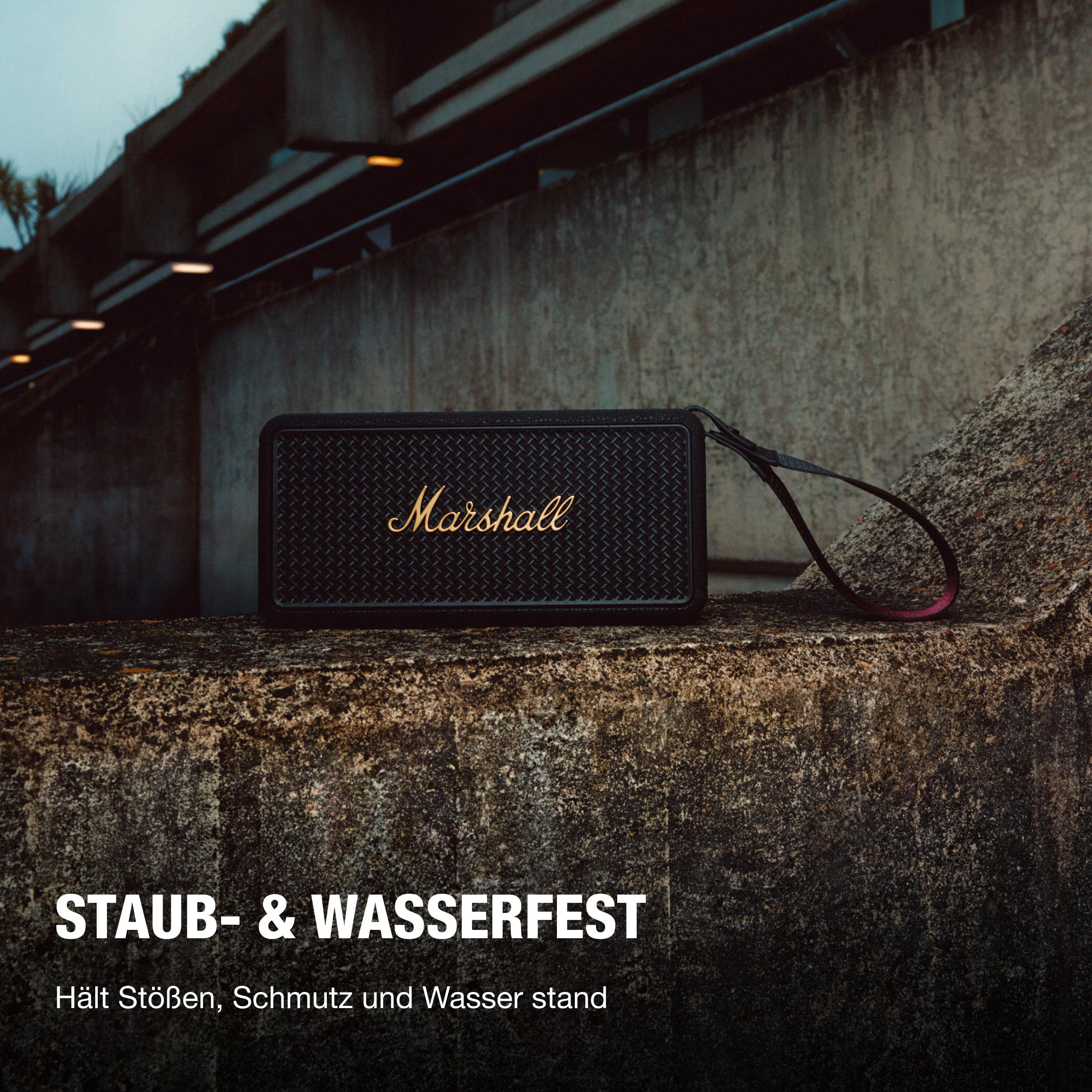 Ein schwarzer Marshall-Lautsprecher auf einer Betonoberfläche. Er hat einen Riemen und den Markennamen in Gold. Hintergrund ist eine Betonmauer.