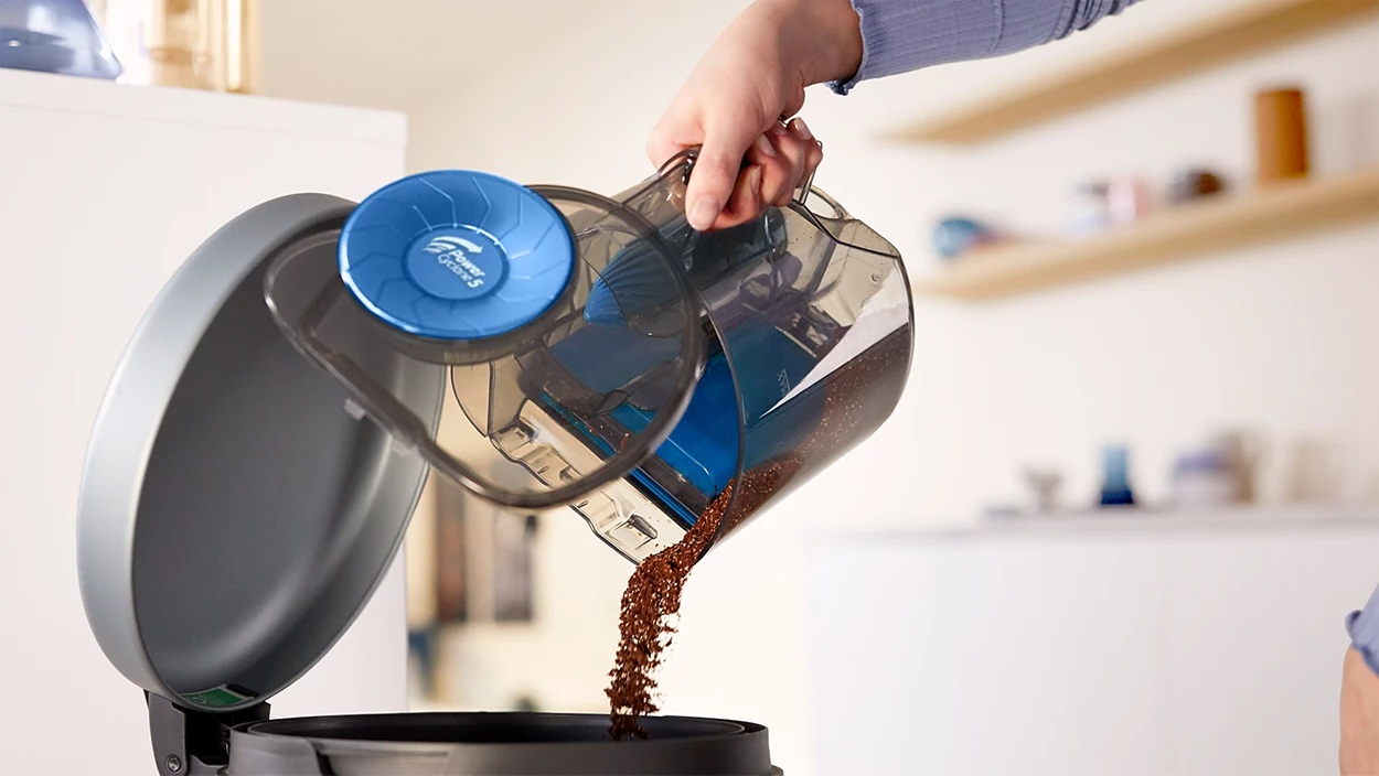 Osoba opróżnia pojemnik odkurzacza do pojemnika. Pojemnik jest przezroczysty, a zawartość to brązowe zanieczyszczenia.