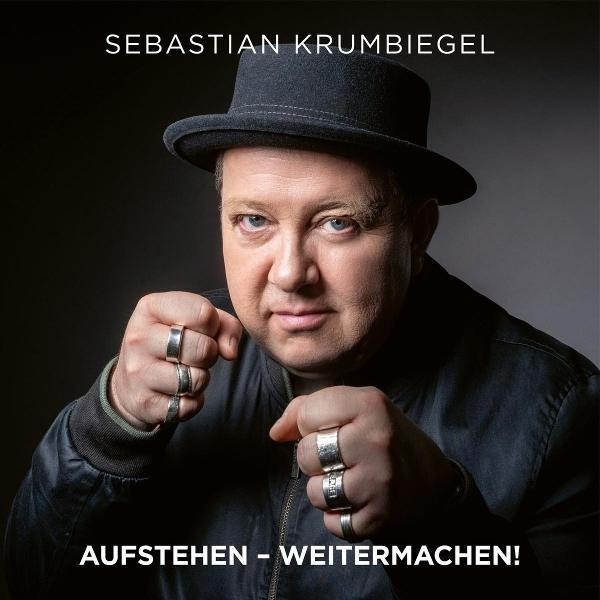 Mann mit Hut und Jacke. Geballte Fäuste, Ringe an den Fingern. Text: Sebastian Krumbiegel, Aufstehen - Weitermachen!