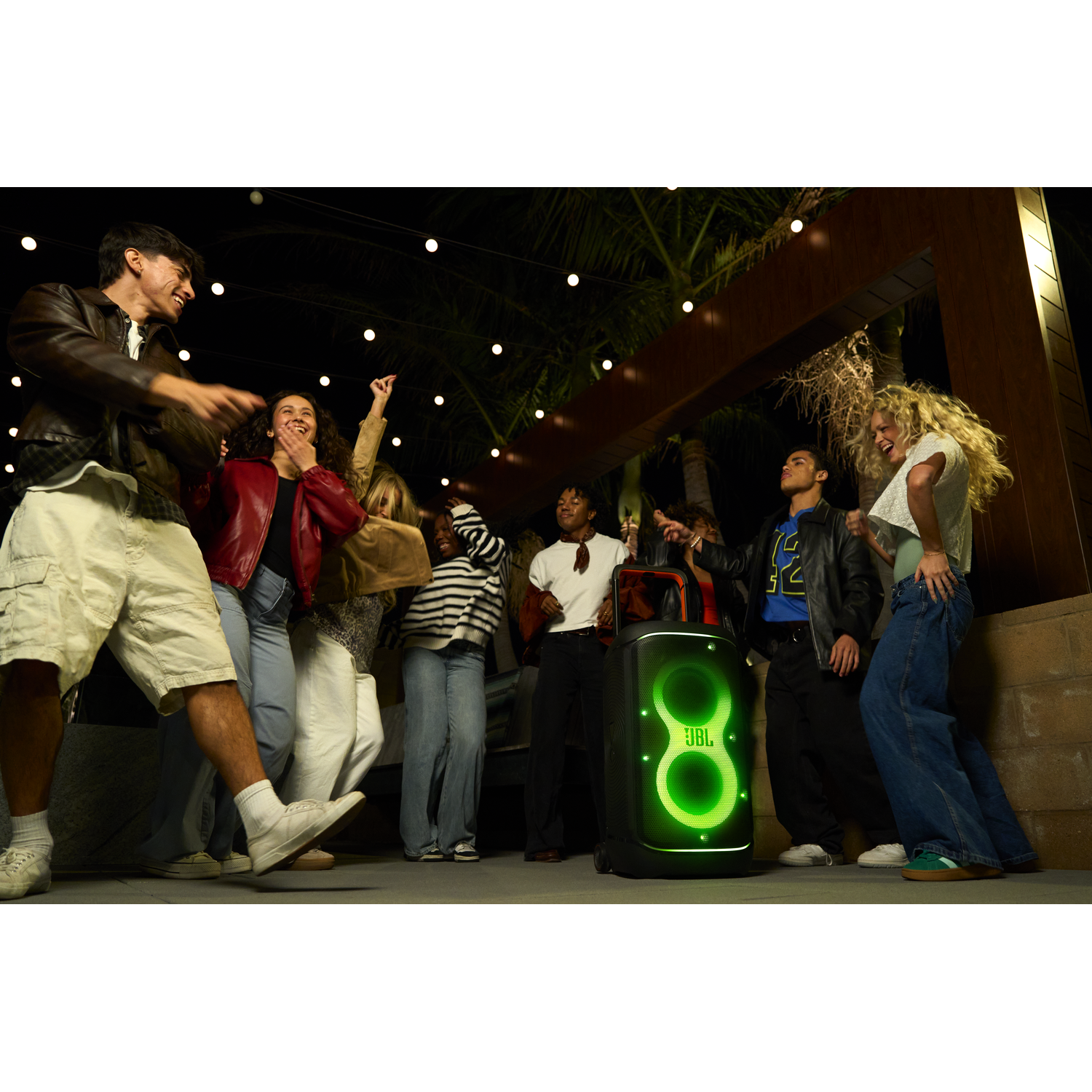 Een groep jonge mensen danst 's nachts in de buurt van een JBL-luidspreker. De luidspreker gloeit groen. Slingers en palmbomen zijn zichtbaar.