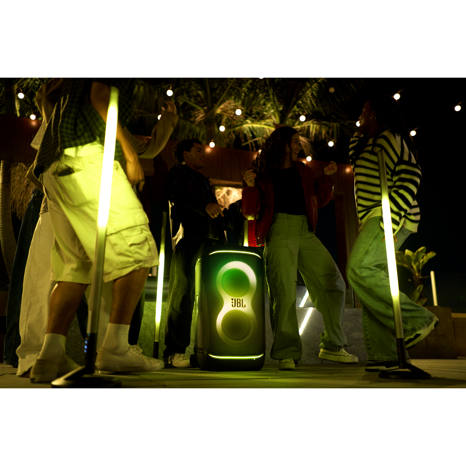 Mensen dansen 's nachts met een JBL-luidspreker. Groene lichten verlichten de scène. Palmbomen en lichtslingers op de achtergrond.