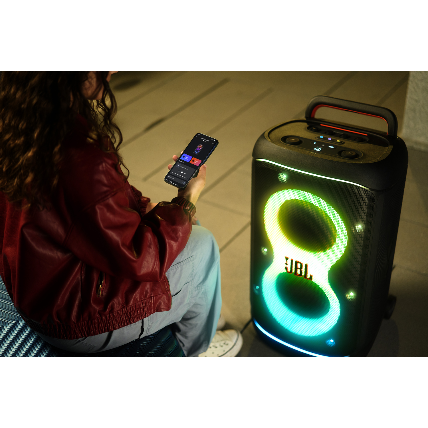 Een persoon gebruikt een telefoon in de buurt van een JBL-luidspreker. De luidspreker heeft kleurrijke lichten. De persoon draagt een rode jas en een blauwe broek.
