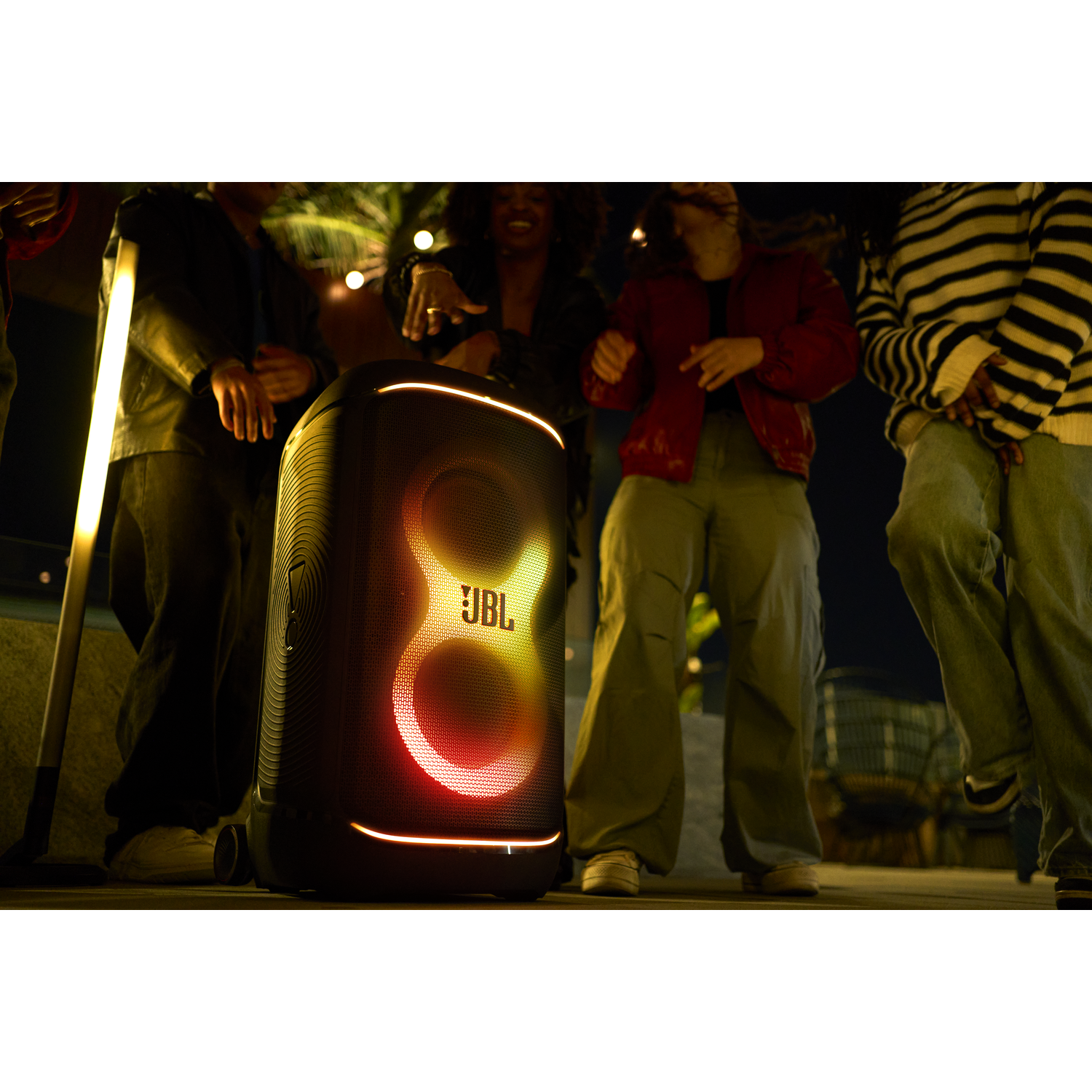 Mensen dansen in de buurt van een JBL-luidspreker met kleurrijke lichten. De scène speelt zich 's nachts af.