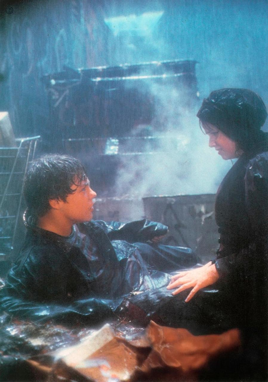 Zwei Personen in schwarzer Kleidung stehen im Regen. Einer liegt, der andere berührt ihn sanft. Im Hintergrund industrielle Elemente.
