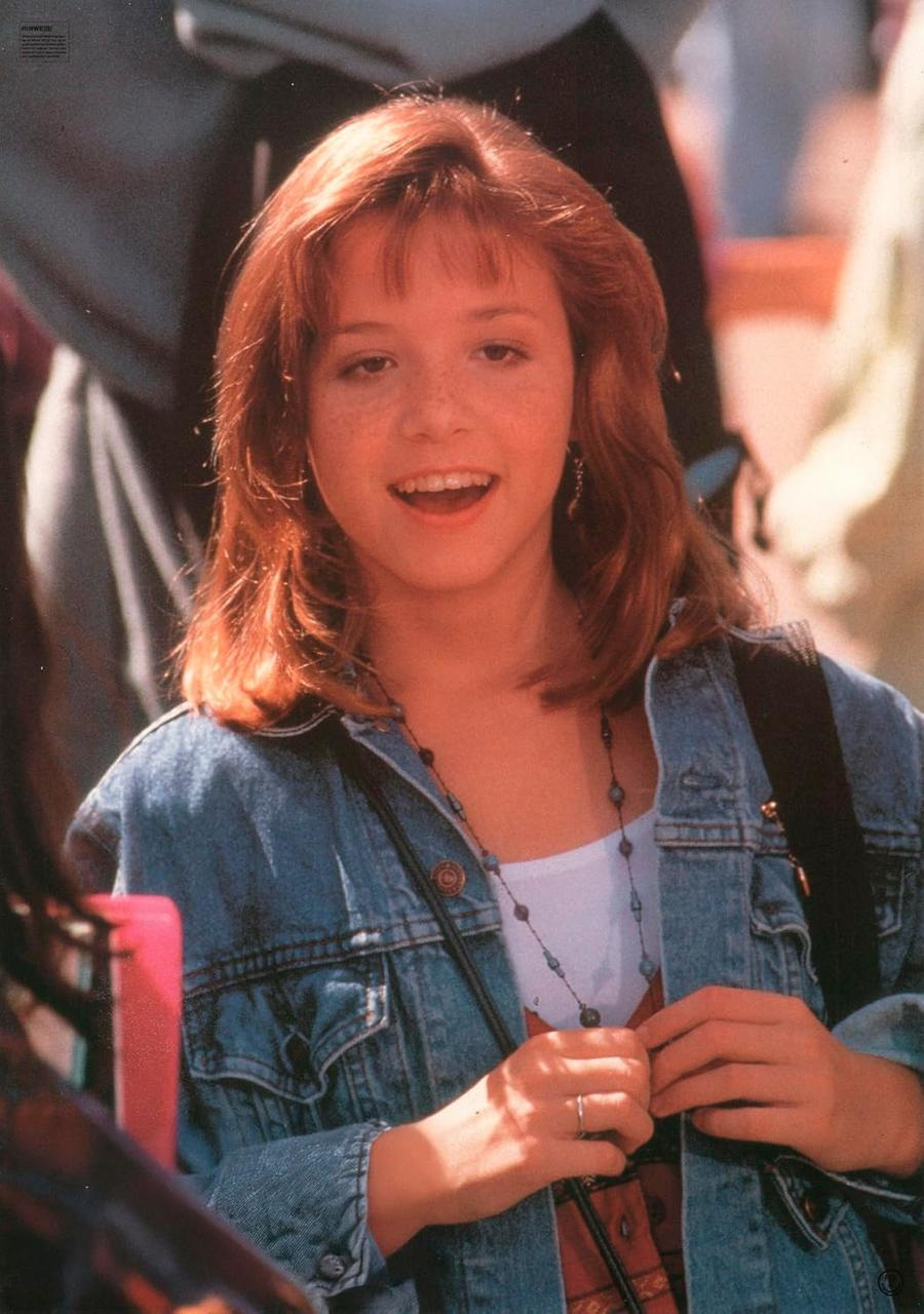 Eine junge Frau mit roten Haaren lächelt und trägt eine Jeansjacke. Sie hat Sommersprossen und eine Halskette. Andere Leute sind im Hintergrund.
