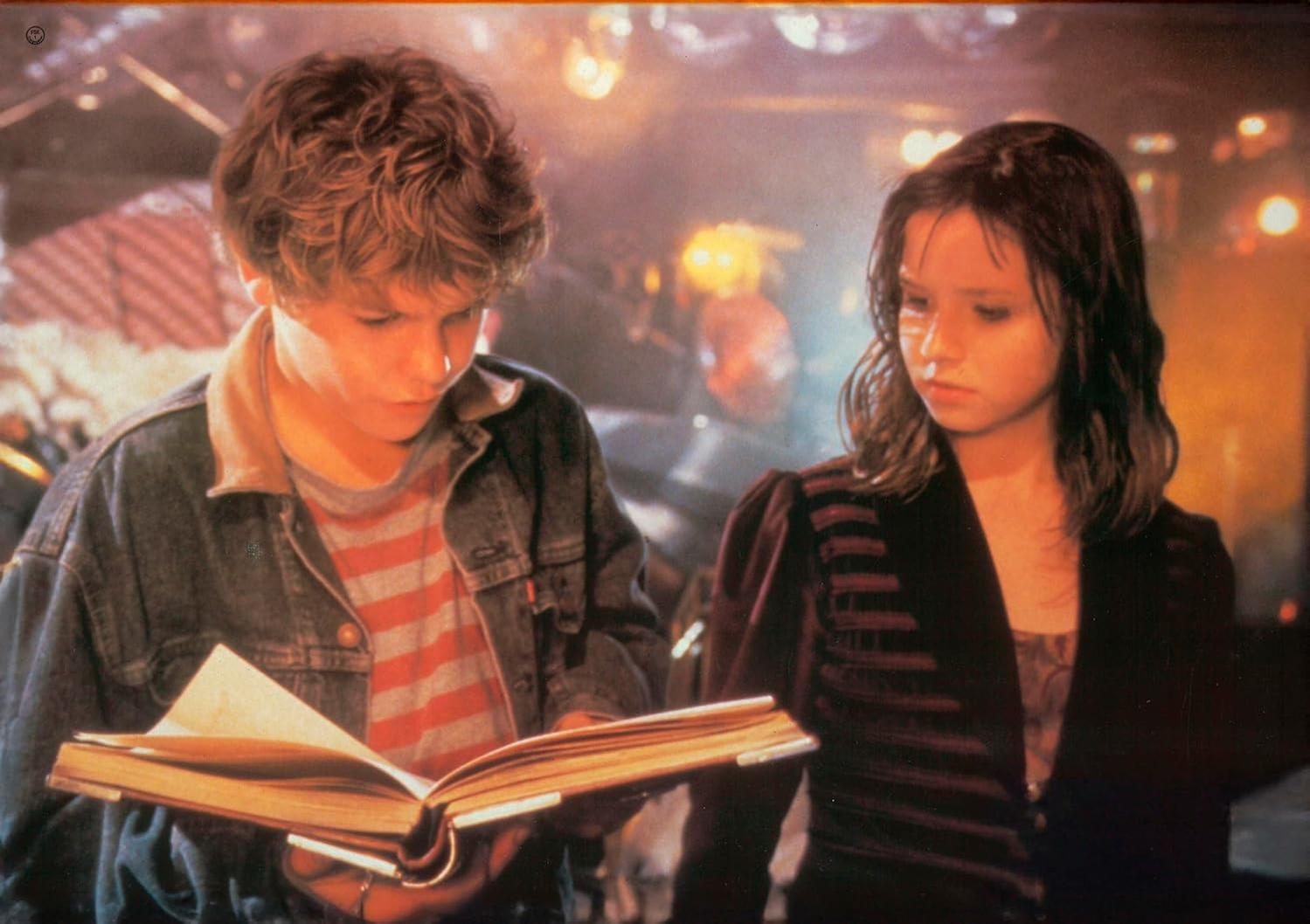 Zwei junge Leute lesen gemeinsam ein Buch. Der Junge trägt eine Jeansjacke und ein gestreiftes Hemd. Das Mädchen trägt eine dunkle Jacke.