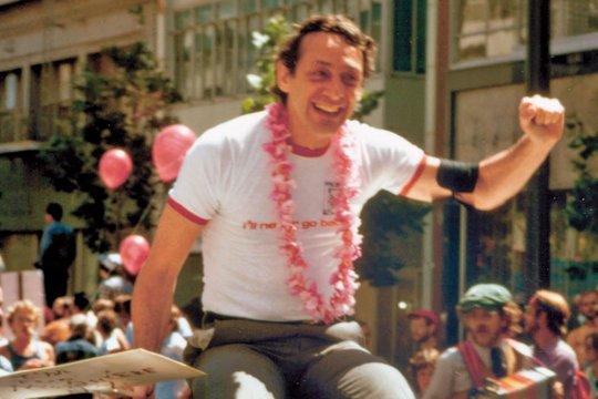 Mann in weißem Hemd mit rotem Saum und Lei hebt die Faust. Rosa Ballons und Menschenmenge im Hintergrund. Lächelt.