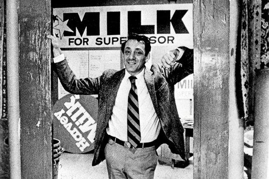 Ein Mann in Anzug, Krawatte und Jacke steht in der Tür. Auf dem Schild steht 'MILK for Supervisor'. Ein rundes Schild zeigt 'Harvey Milk'.