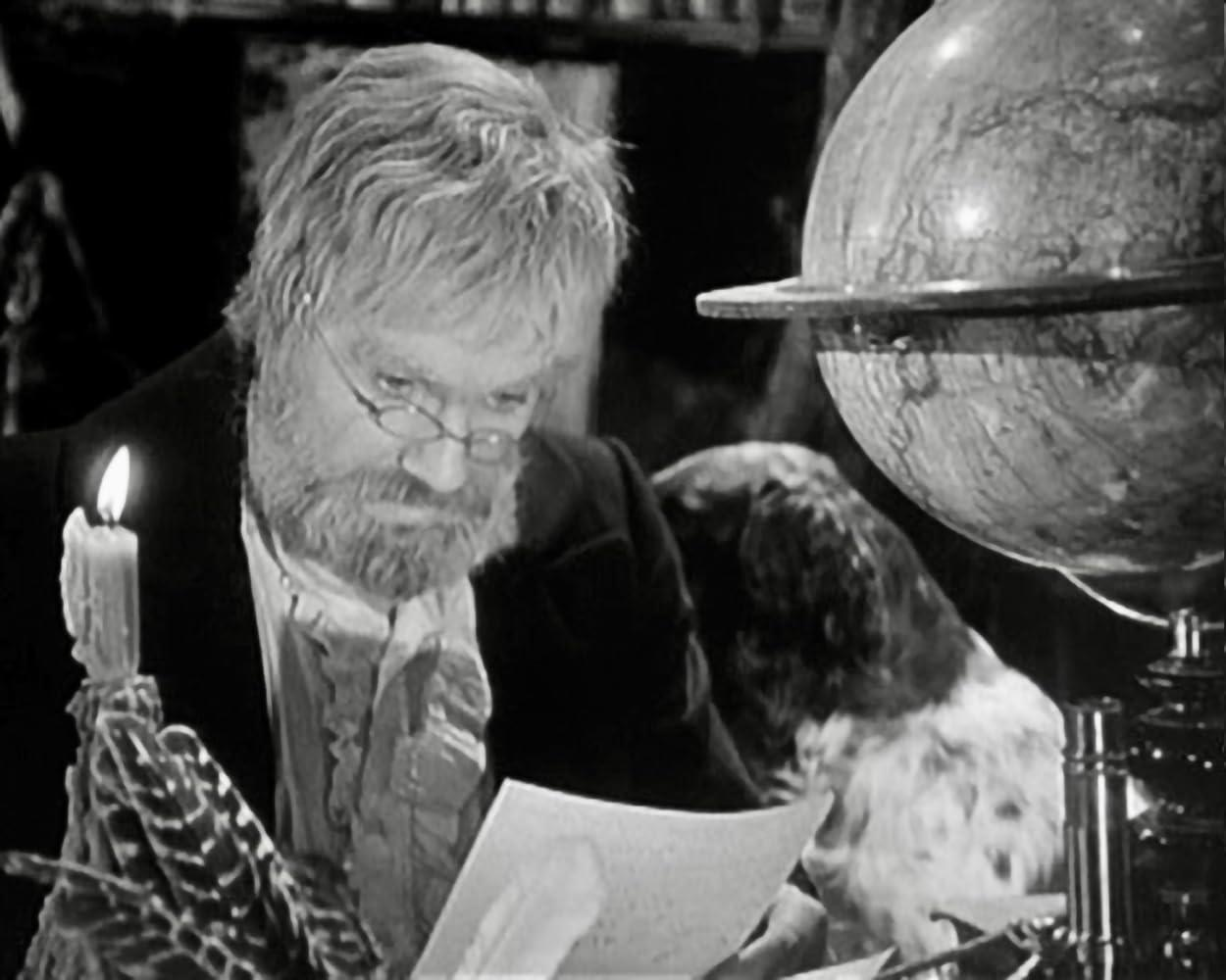 Ein alter Mann mit Bart und Brille liest bei Kerzenlicht ein Papier, im Hintergrund sind ein Globus und ein Hund zu sehen.