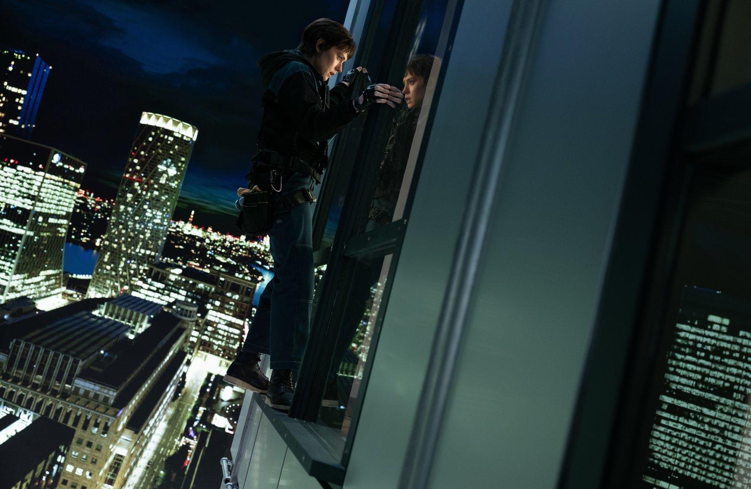 Eine Person steht am Rand eines Gebäudes und betrachtet ihr Spiegelbild im Glas. Stadtlichter beleuchten den Hintergrund bei Nacht.