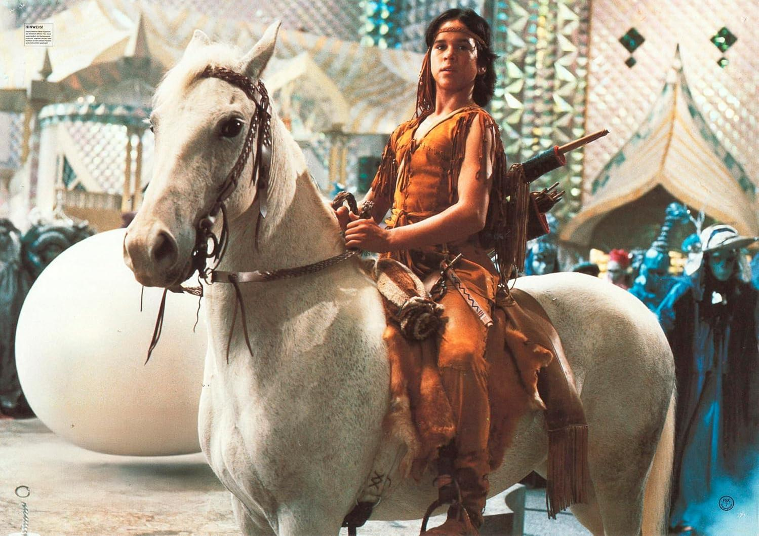 Eine Person in indianischer Kleidung reitet ein weißes Pferd. Im Hintergrund eine große weiße Kugel und kostümierte Figuren.