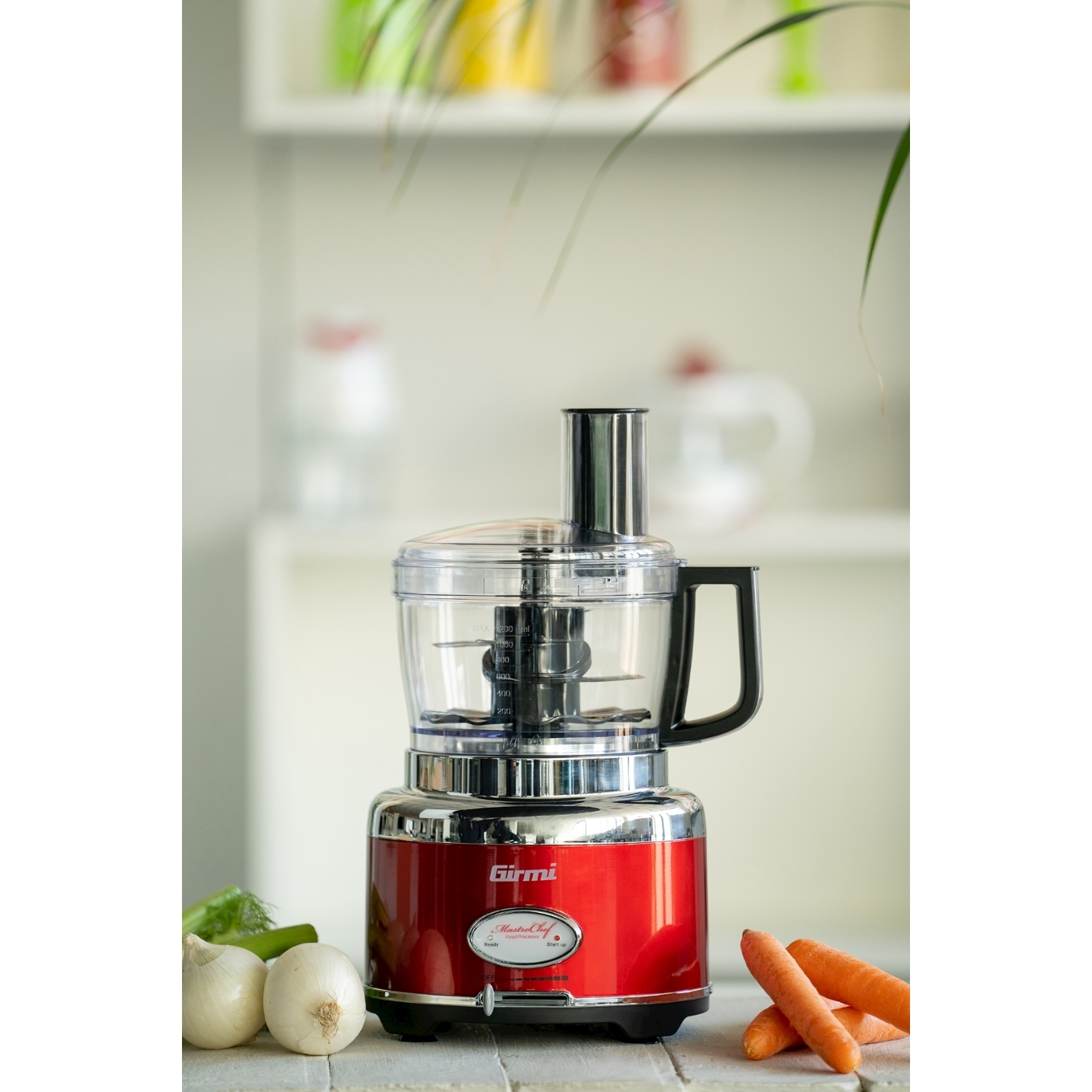 Un robot da cucina rosso con manico nero, cipolle, carote e aneto in primo piano. Lo sfondo presenta piante e scaffali.