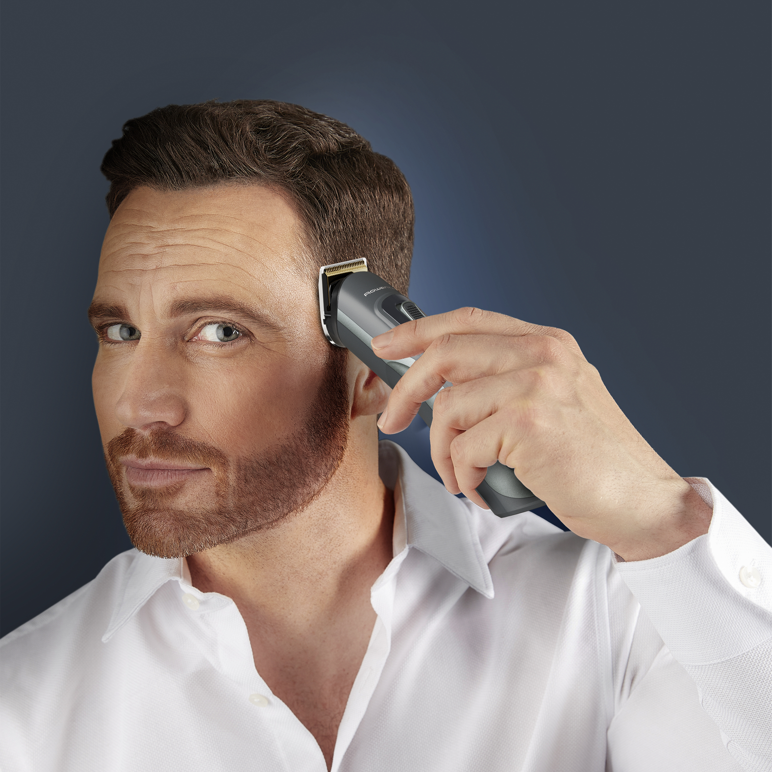 Un hombre se afeita la cabeza con una recortadora eléctrica. Tiene barba y viste camisa blanca.