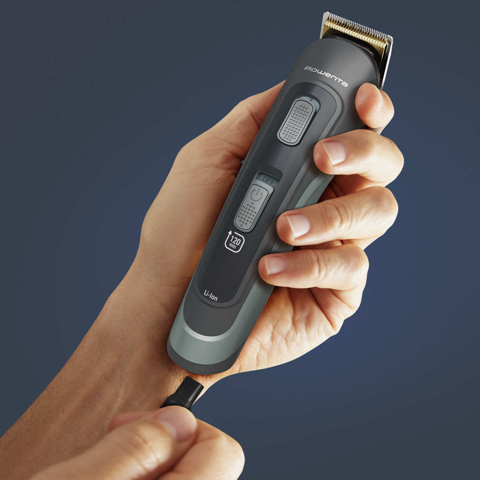 Una mano sostiene una recortadora de cabello Rowenta, conectando un cable negro. La recortadora es gris y negra, con botones de control.