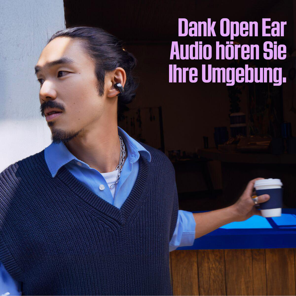 Mann mit Ohrhörern hält Kaffee. Er trägt ein blaues Hemd, einen marineblauen Pullover und eine silberne Kette. Text: 'Dank Open Ear Audio'.