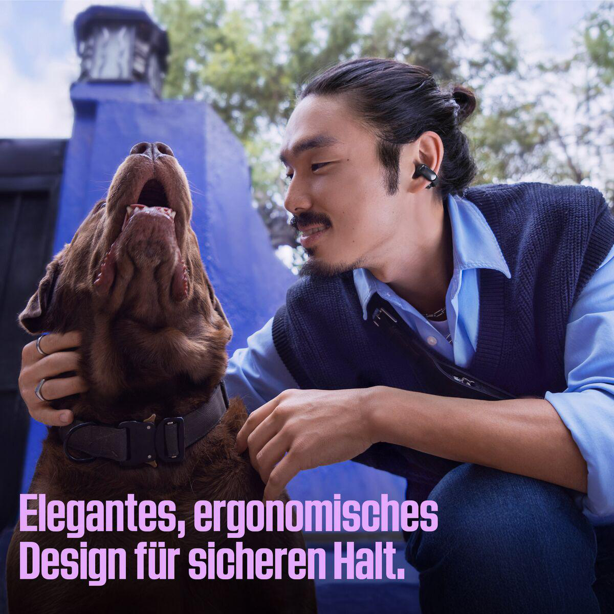 Mann und Hund. Mann mit Ohrhörern, streichelt einen braunen Hund. Hintergrund ist eine blaue Wand und Laub.