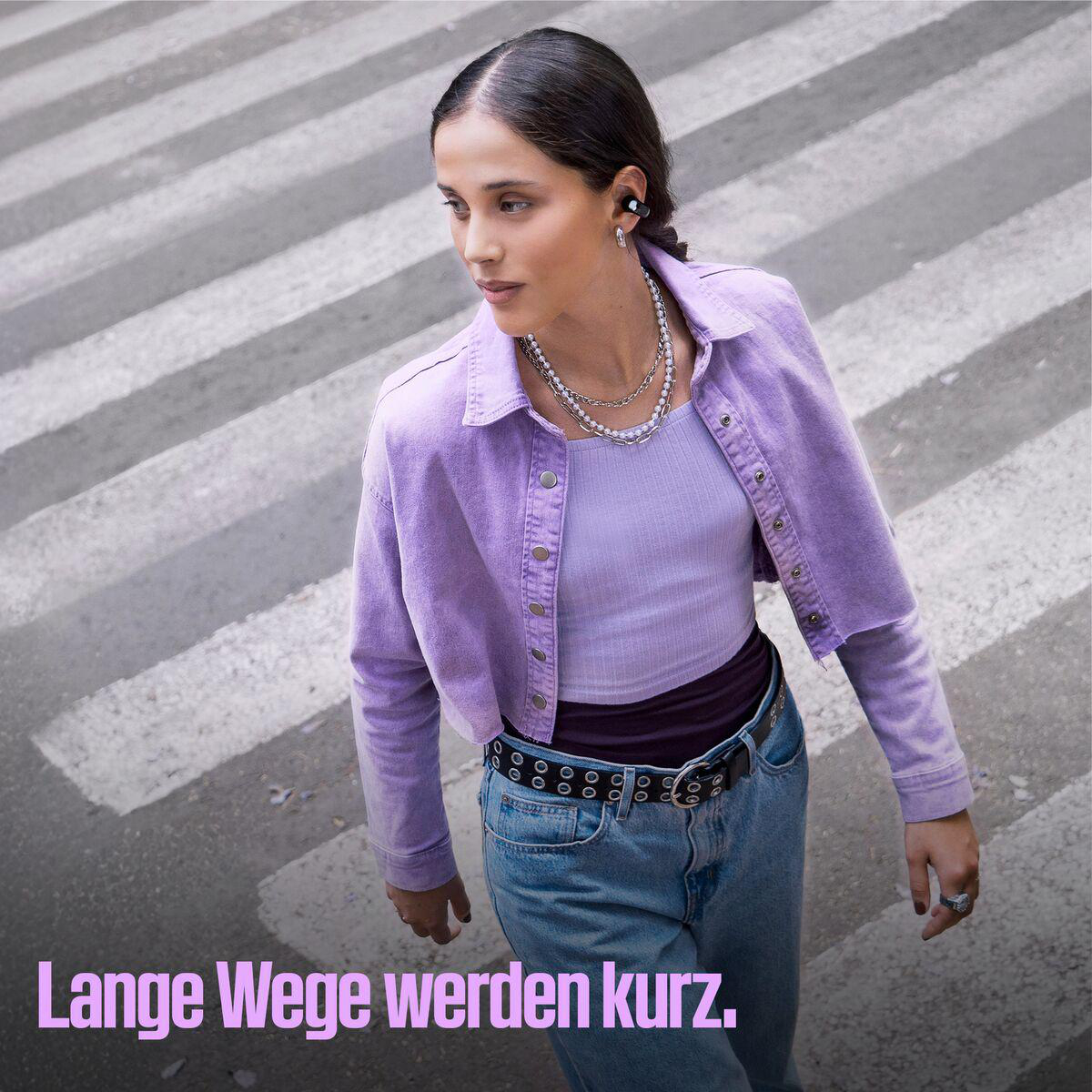 Frau in lila Jacke und Jeans mit Ohrhörern auf einem Zebrastreifen. Text: 'Lange Wege werden kurz'.
