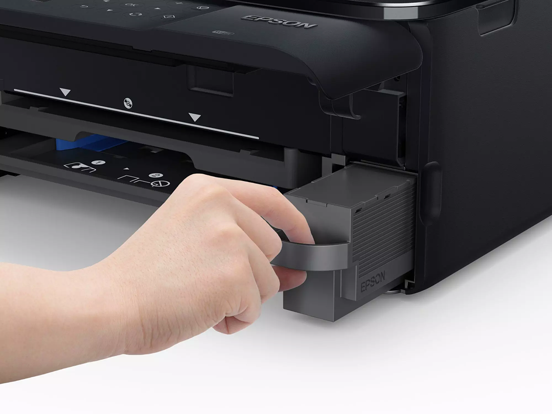 Egy kéz kihúz egy szürke tintapatront egy fekete Epson nyomtatóból. A nyomtató fehér felületen van.