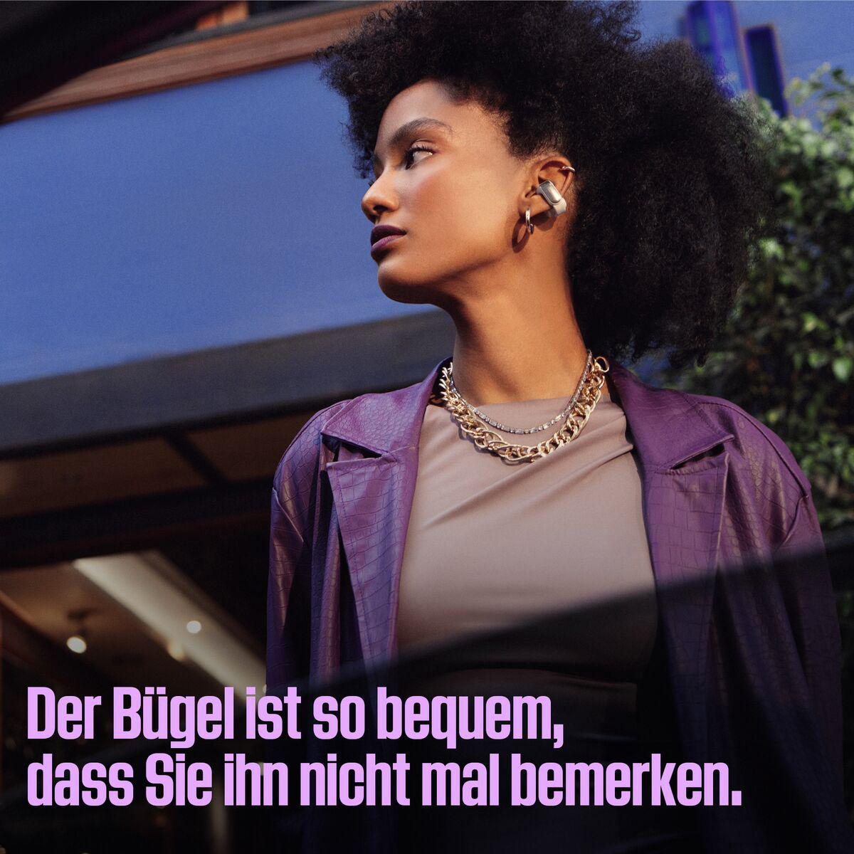 Eine Frau mit lockigem Haar trägt eine lila Jacke und Goldschmuck. Sie hat Kopfhörer im Ohr. Der Hintergrund hat blaue und grüne Elemente.