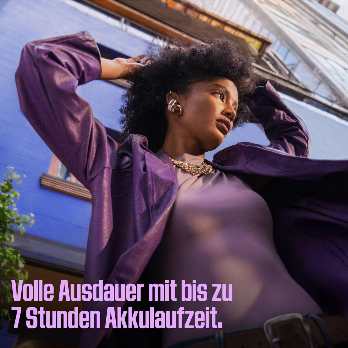 Frau in Lila mit lockigem Haar, trägt Goldschmuck und eine Jacke. Im Hintergrund ein blaues Gebäude und Text: „Volle Ausdauer“.