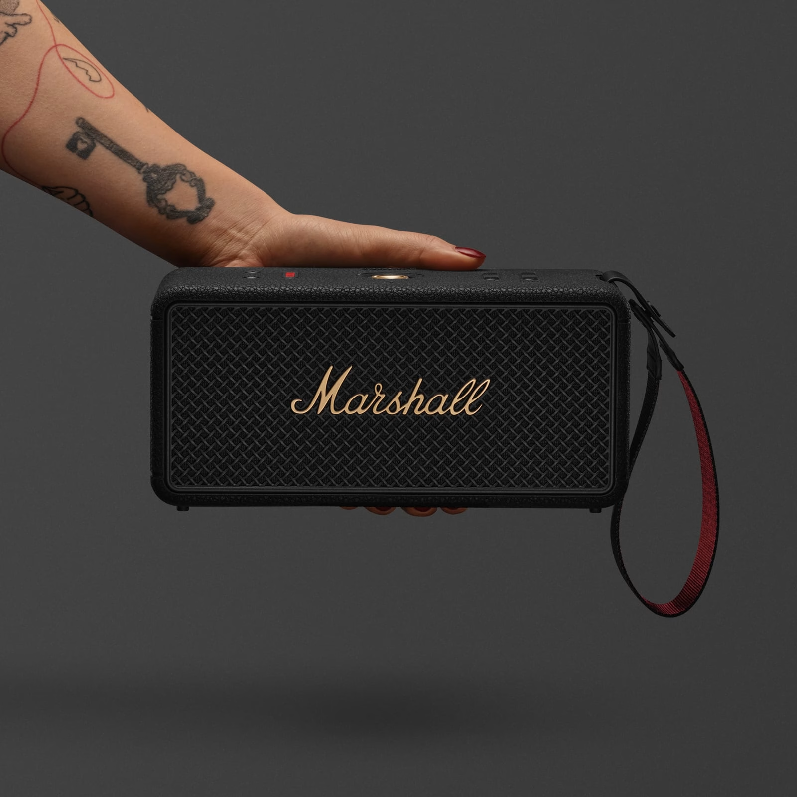 Una mano tiene un altoparlante Marshall nero. Un tatuaggio a chiave è visibile. L'altoparlante ha un logo dorato e un cinturino rosso.
