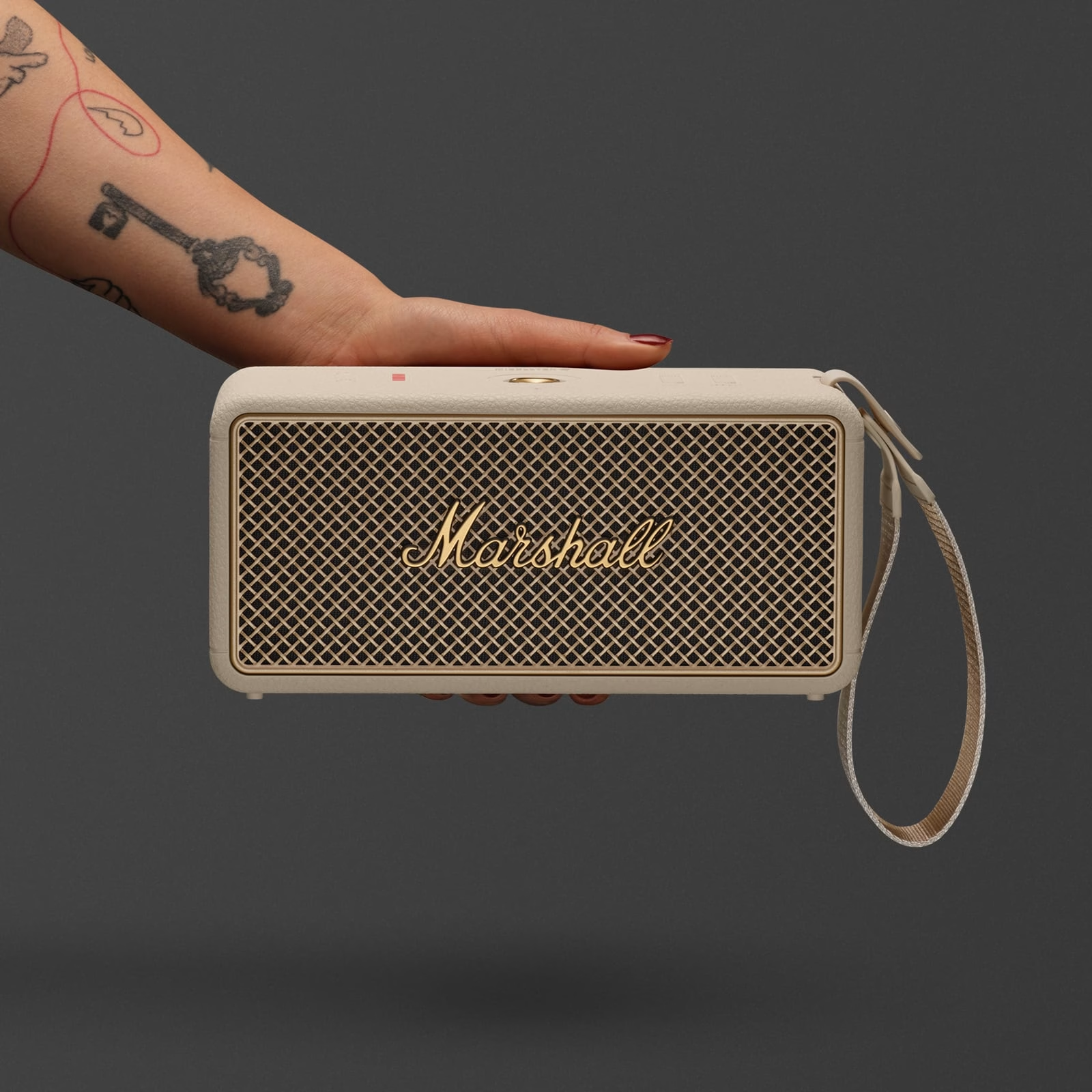 Eine Hand hält einen cremefarbenen Marshall-Lautsprecher. Der Lautsprecher hat eine goldfarbene Gitterfront und das Wort "Marshall".