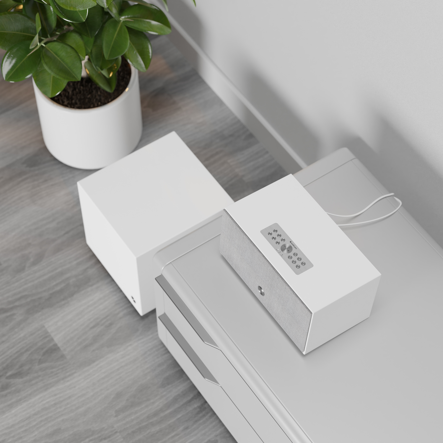 Een witte luidspreker met afstandsbediening op een grijze kast, naast een witte doos en een plant in een witte pot. Grijze houten vloer.
