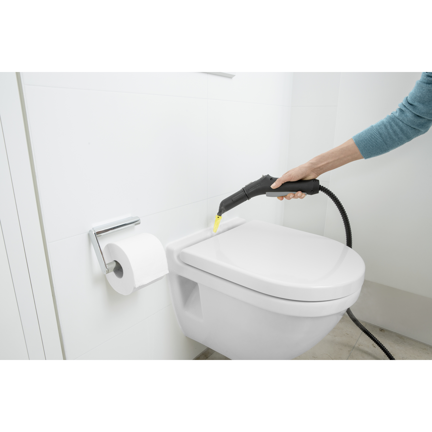 A person uses a steam cleaner on a white toilet. A roll of toilet paper is on the left. The wall is white.