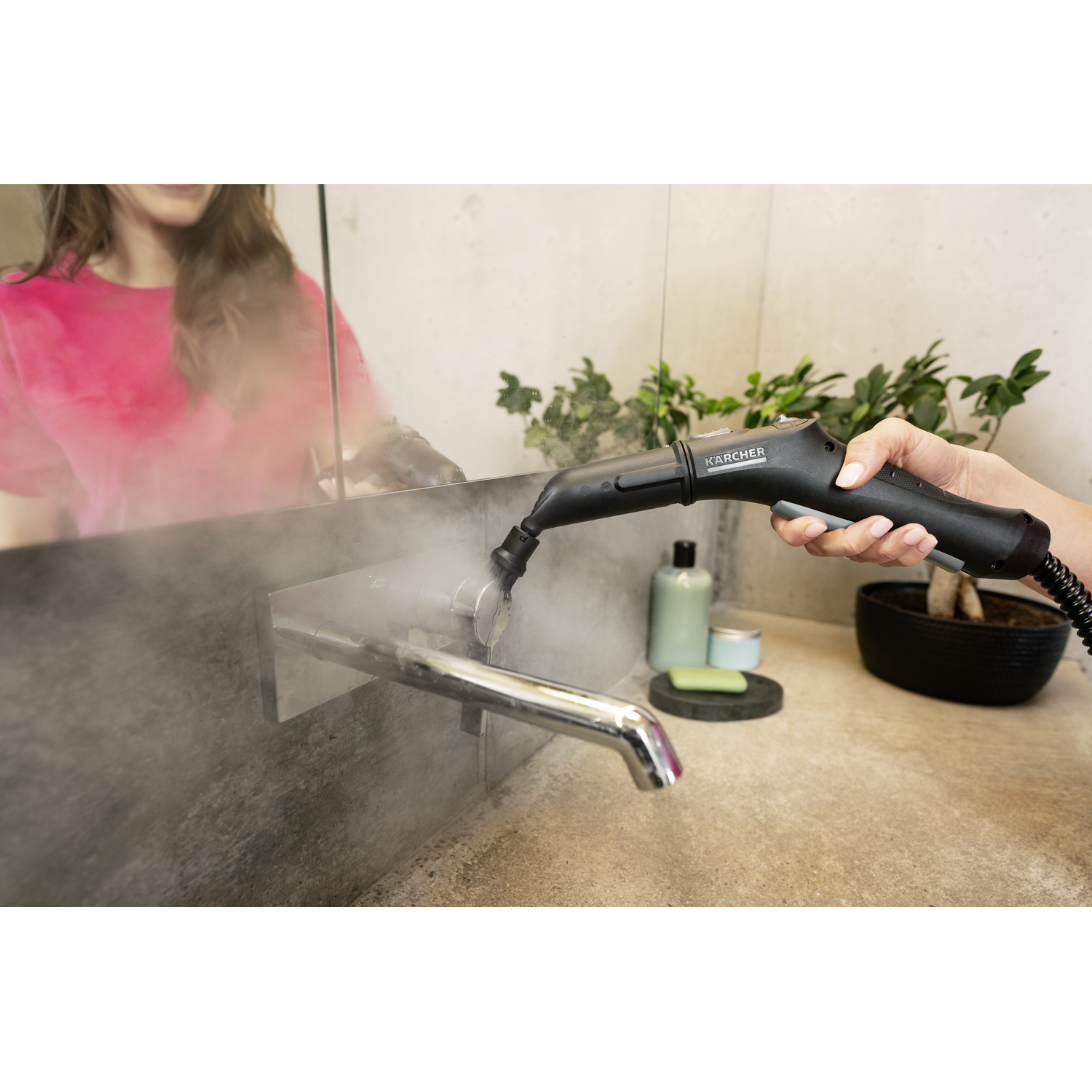A person using a steam cleaner on a bathroom faucet. Steam is emitted. A mirror reflects the person and a pink shirt.