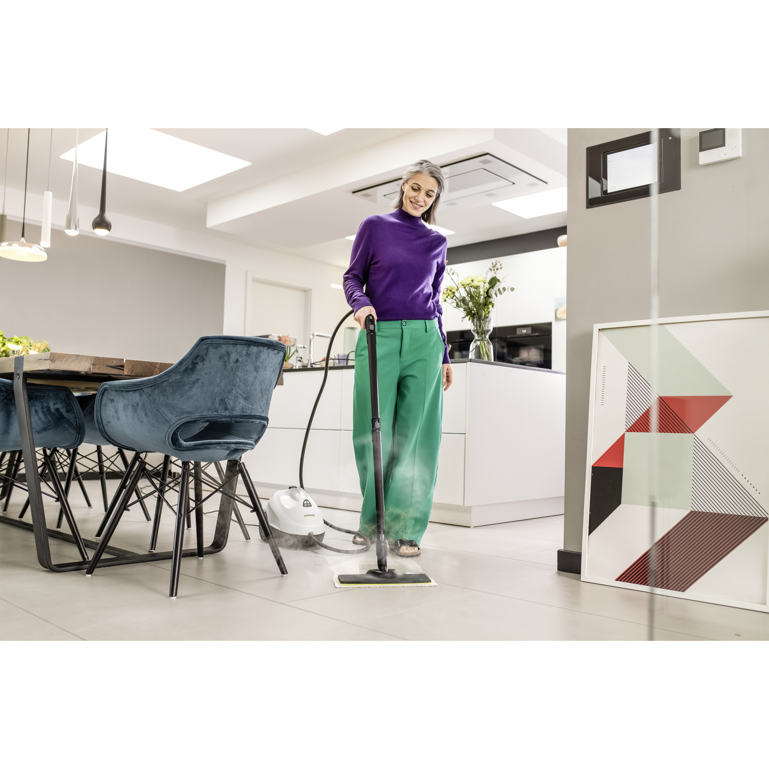 Woman in purple top and green pants uses a steam cleaner on a tile floor. Kitchen and dining area in background.