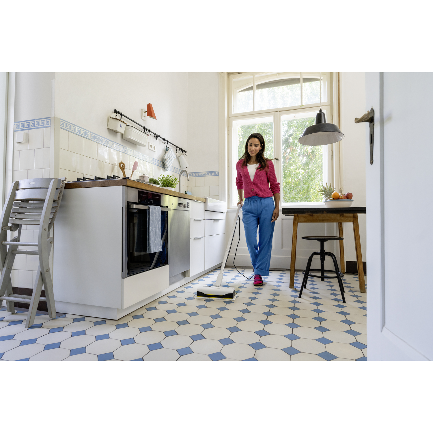 Woman in pink top and blue pants mops a kitchen floor with geometric tiles. Background includes a window and table with fruit.