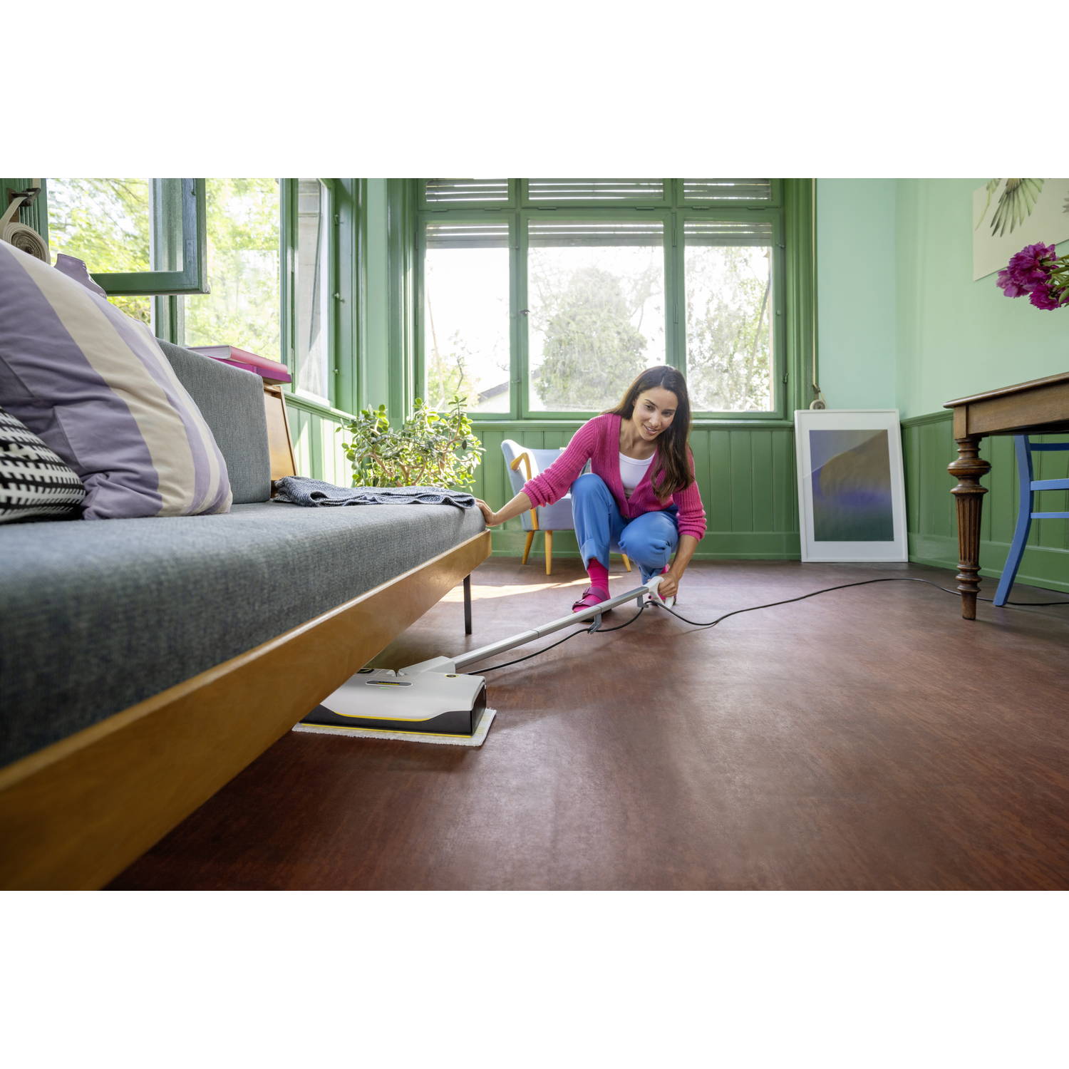 A woman in pink top and blue pants cleans a wooden floor with a steam cleaner. A sofa and window are in the background.