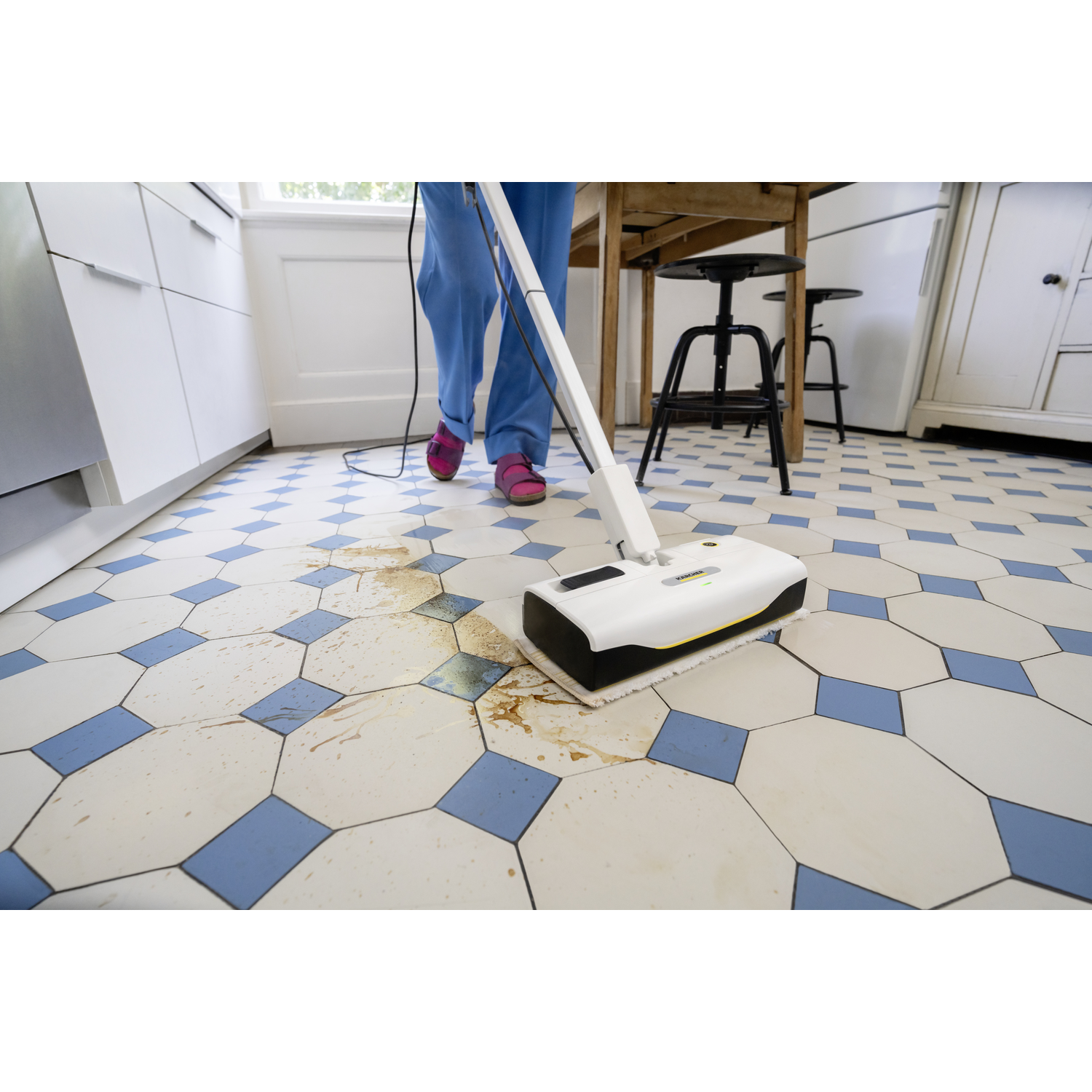 A person in blue pants and pink shoes is cleaning a tiled floor with a white and black cleaning device. The floor is dirty.