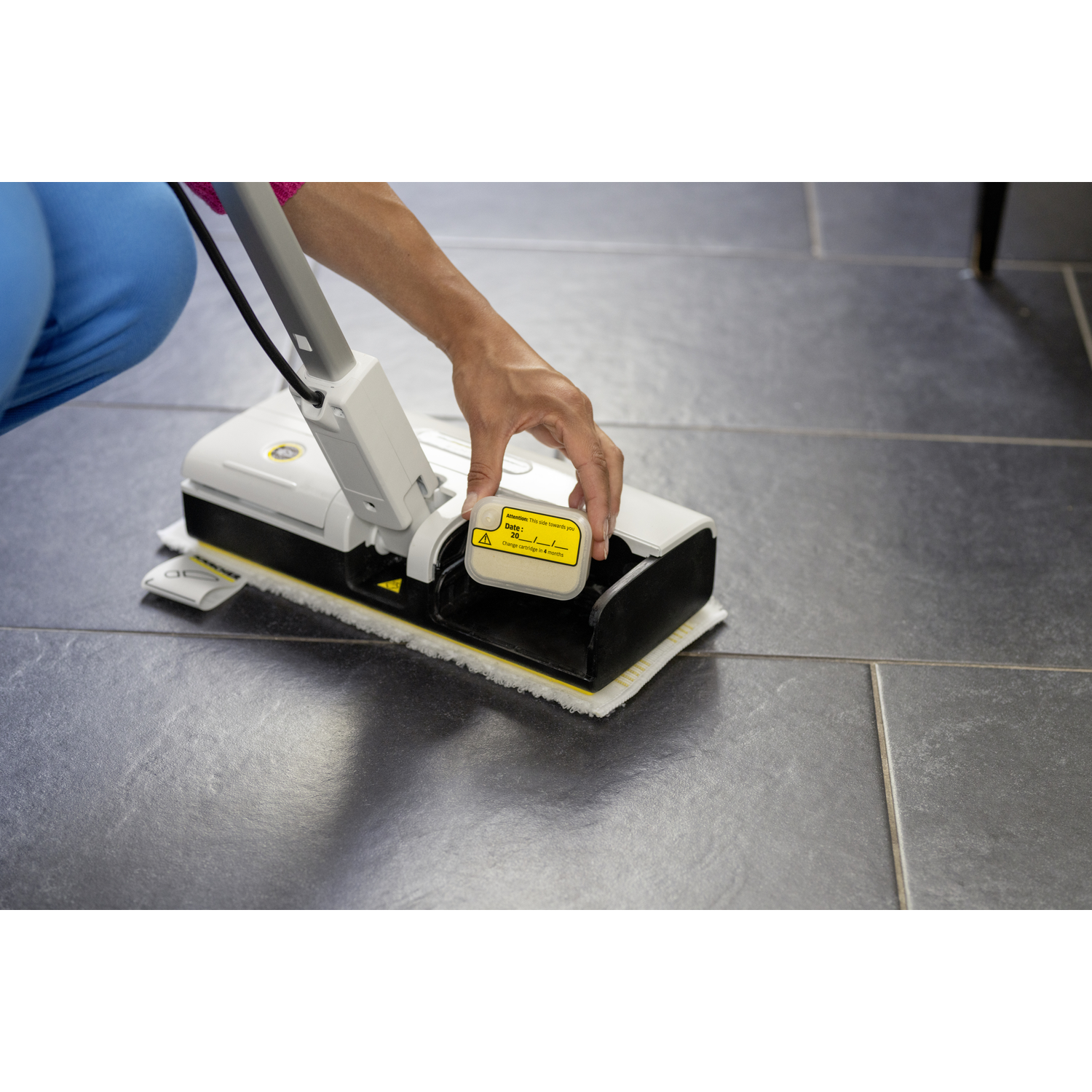 A person uses a white and black floor cleaner on gray tiles. A yellow cartridge is being inserted into the cleaner.