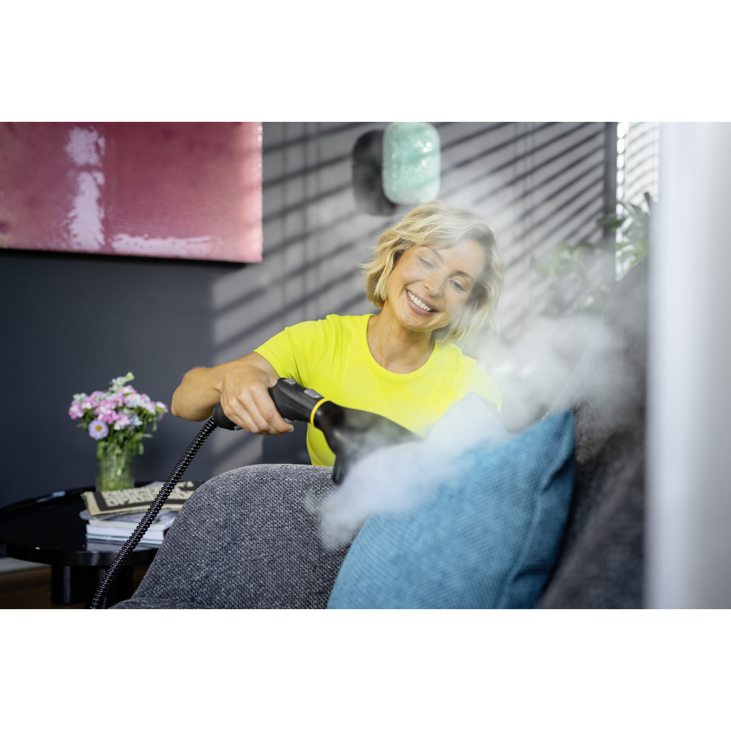 Une femme nettoie un canapé avec un nettoyeur vapeur. Elle porte une chemise jaune et sourit. De la vapeur s'élève du coussin bleu.
