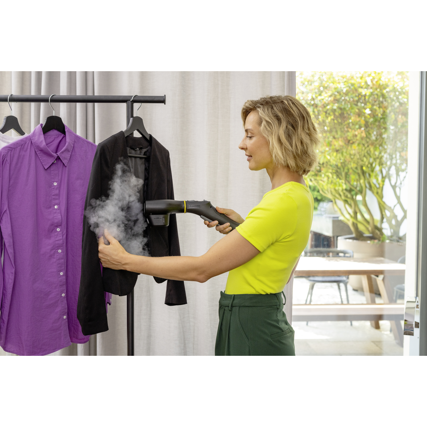 Une femme repasse une veste noire sur un portant. Des chemises violettes et blanches sont accrochées à côté. Elle porte un haut jaune et un pantalon vert.