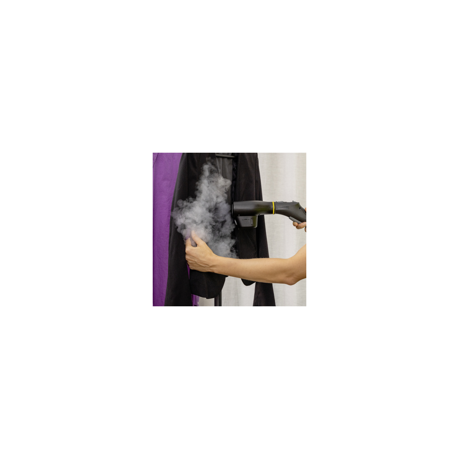 Une personne qui repasse une veste noire avec un nettoyeur vapeur. De la vapeur est visible. Tissu violet à gauche, tissu blanc derrière la veste.